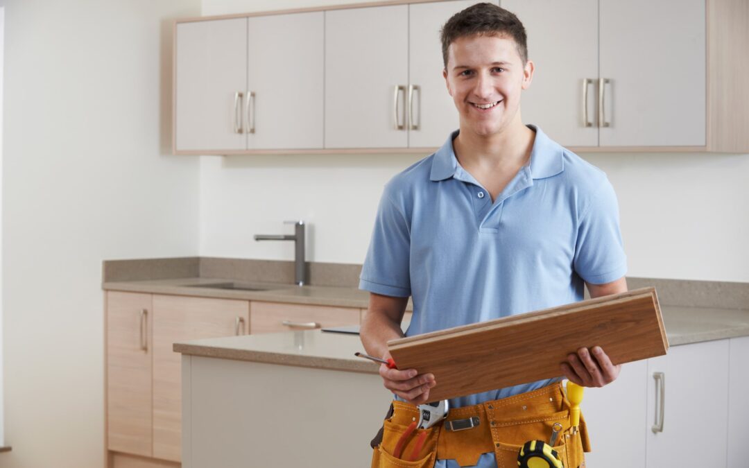 A contracting professional standing in a recently remodeled kitchen, holding a piece of wood flooring and wearing a toolbelt